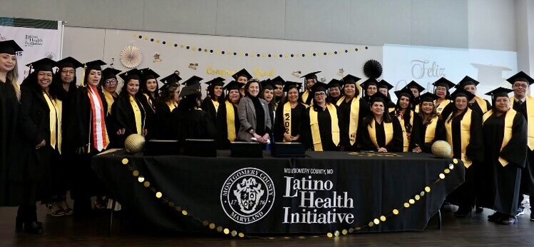 Graduates pose with Council President Stewart in front of a black and white Latino Health Initiative banner.