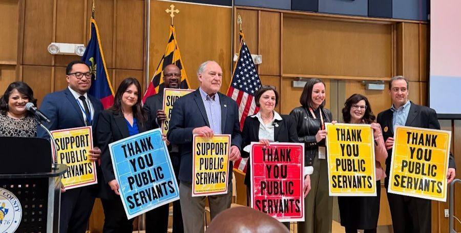 Elrich, Stewart and county leaders stand on a stage holding signs reading “Support public servants” and “Thank you public servants”.