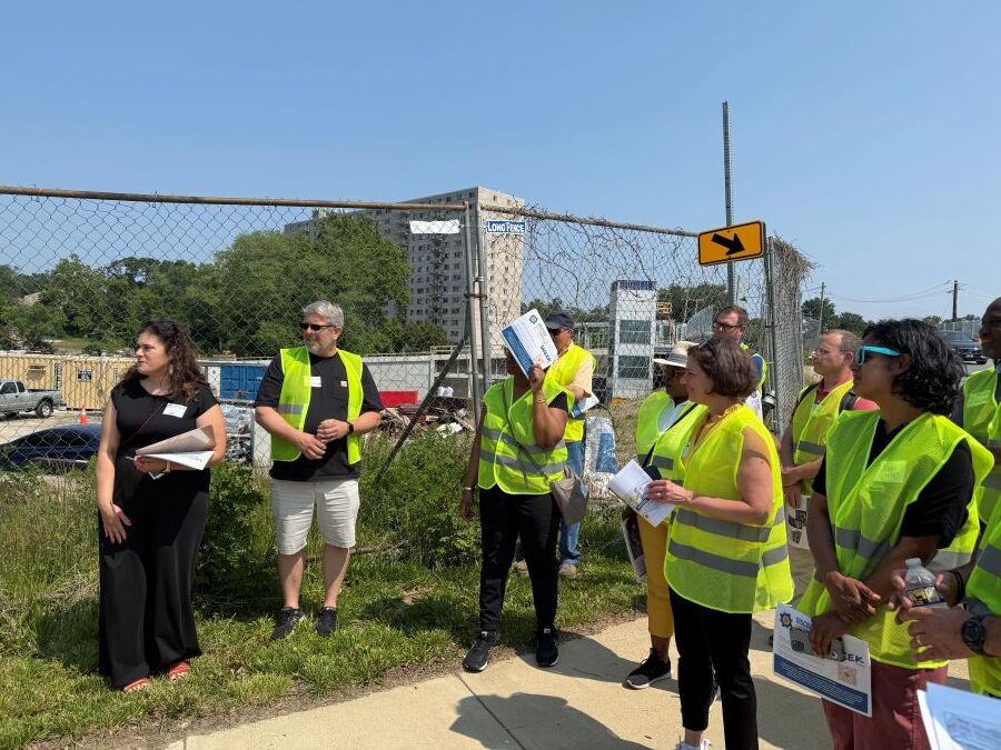Community members stand on a sidewalk wearing yellow high-visibility vests during the Lyttonsville community walk. 