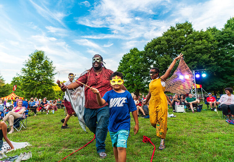 Adult and two children with colorful masks and butterfly wings at a park festival.