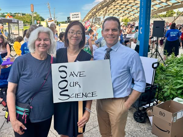 Councilmember Glass and two women hold a sign that reads "Save Our Science"