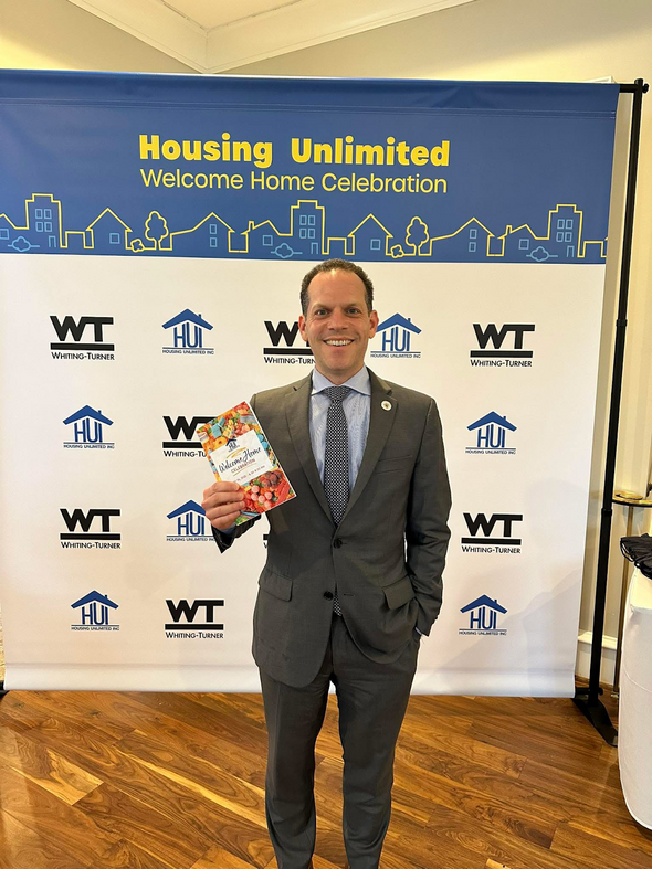 Councilmember Glass in front of a banner that reads "Housing Unlimited Welcome Home Celebration"