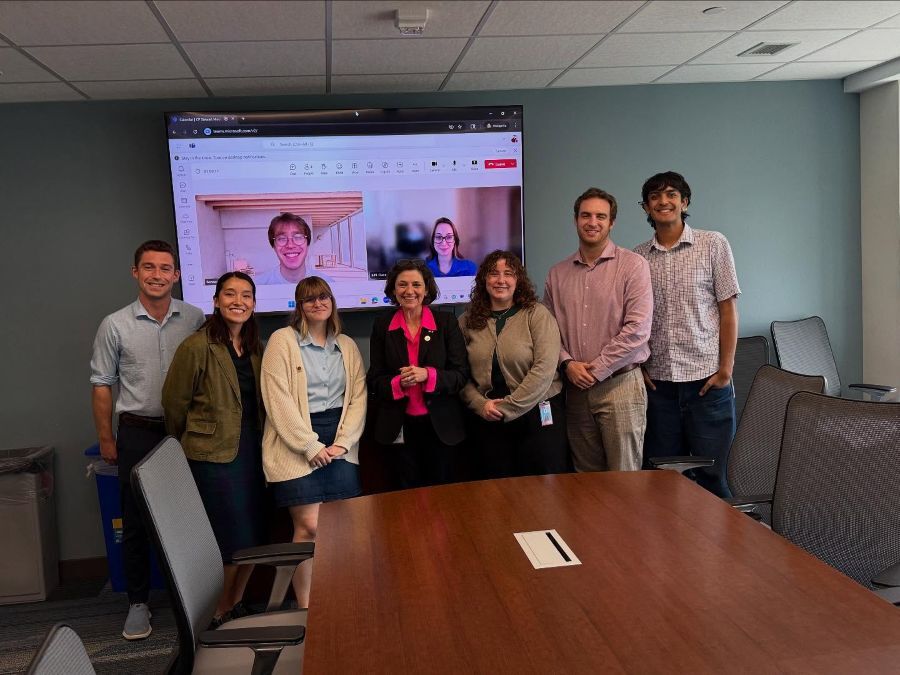 Council President Stewart and Council Fellows pose in a conference room. Two people join virtually on a Zoom screen in the background.