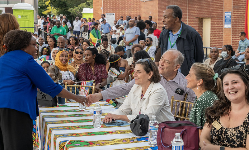 Council President Stewart sits at a table with elected officials at the Ethiopian Day Festival with a large crowd in the background.