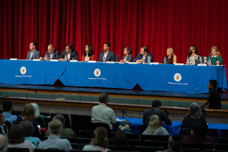 Councilmembers sit at a long table on an auditorium stage in front of an audience.