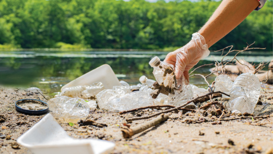 hand picking up water bottle and litter on the ground in front of a body of water