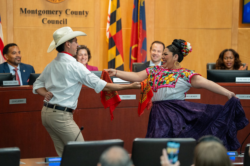 Two folkloric dancers in the Council chamber with Councilmembers smiling in the background.