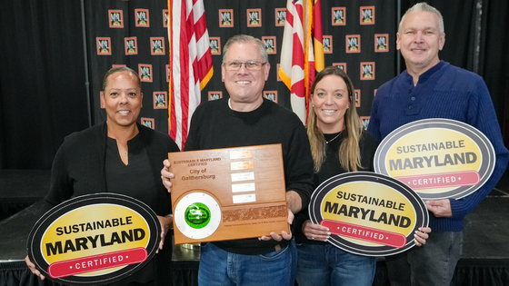 Four individuals proudly displaying awards from Sustainable Maryland, including a wooden plaque and two round signs, at an event.