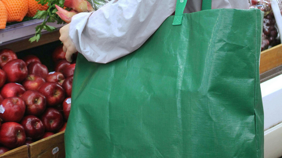 person standing in front of a pile of apples and oranges on a shelf with a green reusable bag on their shoulder.