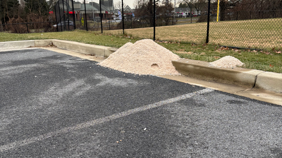 Salt pile sitting on the curb in a parking lot. A shopping center and field is behind the salt pile.