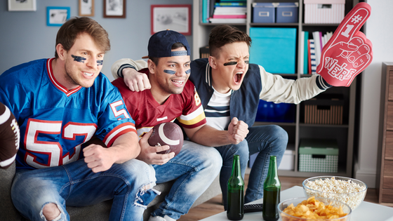Three men sitting on a couch cheering while wearing football jerseys. One man has a foam finger that says "We're number 1."