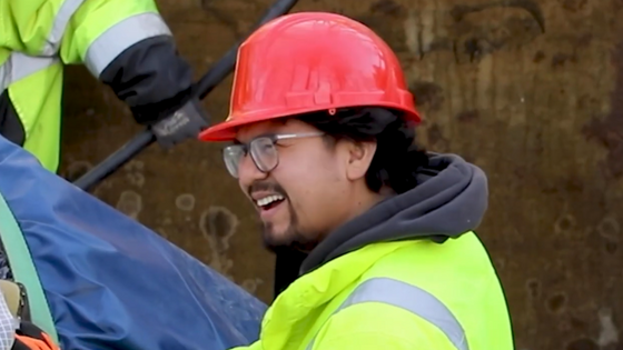 A man in the ground with a vest and hard hat helmet on  smiling