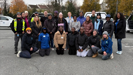 Group of people standing in three rows smiling in a parking lot on a cold, cloudy day.