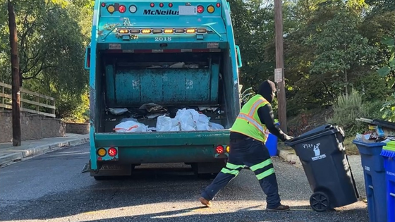 A man loading trash into the back of a garbage truck. The man is dressed in his work uniform