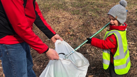 Little boy picking up litter using a grabbing tool and placing it into a trash bag another person is holding open for him.