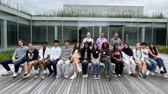 Group shot of people sitting and standing behind a bench smiling in a courtyard on a cloudy day. 