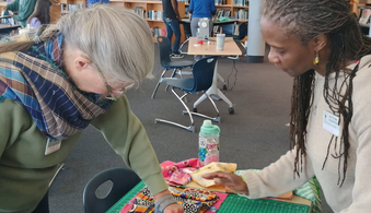 Two women standing on opposites of a table with fabric between them in a library