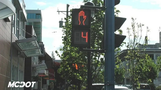 Photo of signal in downtown city with stores and trees in background. Has MCDOT logo.