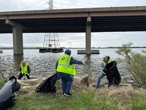 photo of MPA volunteers wearing green construction vests picking up waste along Bear Creek with water and a bridge in the background. 