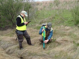 Two people wearing waders and high visibility vests are in a grassy wetland area with equipment used for drone surveys. 