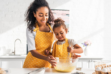 Photo of parent and child whisking and stirring food in a bowl in kitchen wearing aprons.