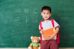 Child holding books and backpack in front of school chalkboard.