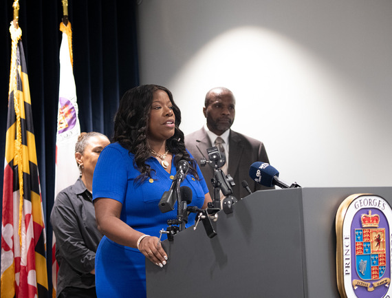 County Executive Braveboy speaking into microphones at podium in front of flags at press conference