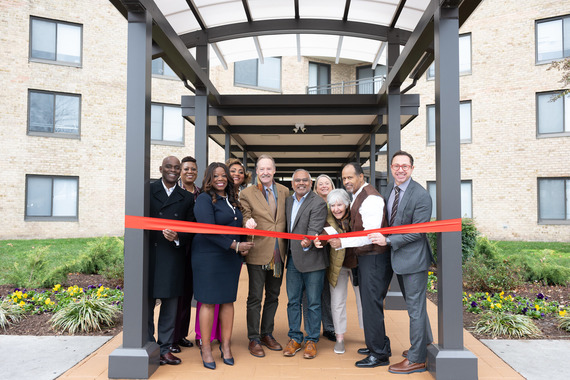 County Executive smiles and holds large scissors with small crowd of people hold red ribbon in front of Plaza Towers