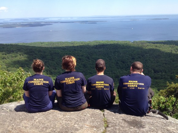 4 Team Members sit on a mountain summit looking out over the ocean.