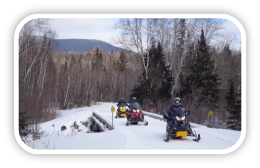 Three snowmobile riders crossing trail bridge with mountain scenery in background.