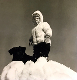 Toddler with black Labrador looking down from atop a snowbank.
