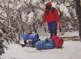 Winter camper with equipment in a snowy forest clearing.