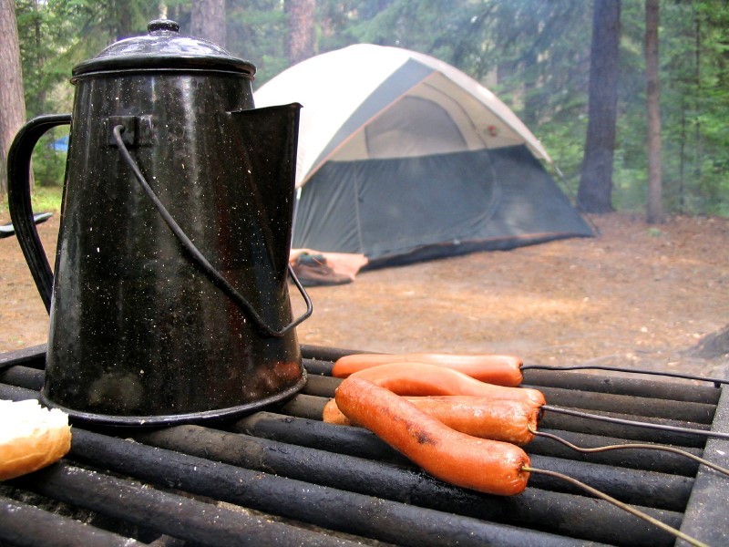 Summertime woodland campsite with coffee and hotdogs on the grill and a dome tent in the background.