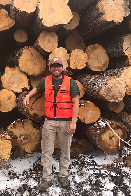 Photo of Jeremy Gignon, new BPL Forestry Technician, in front of a stack of large bole-sized logs.