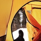 Photo looking out the door of a yellow dome tent across a booted foot to the snow and woods beyond.