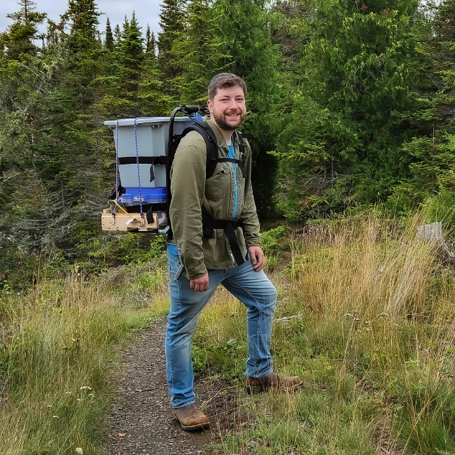 Photo of Riley McManus at a forest edge trail carrying his ceramics bucket on a back frame.