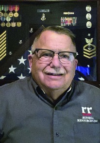 John Hart smiling with military medals and US flag in the background