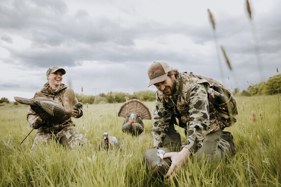 female and male hunters dressed in all camo placing hen turkey decoy while laughing in a field