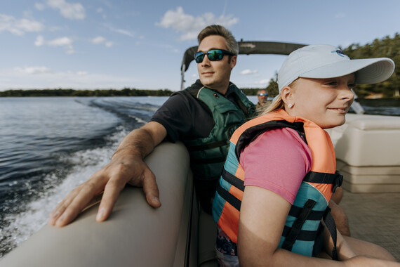 father and daughter riding on pontoon with blue skies above. The daughter is wearing a neon orange and blue lifejacket.