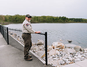 Man standing alone lake shoreline with tarred pathway underneath and a chain-link fence