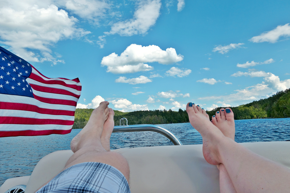 two people laying on back of pontoon boat with bright sunny skies and American flag waving