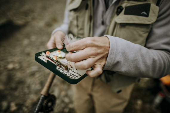 woman picking out a fly from her fly box on a river bank wearing tan waders