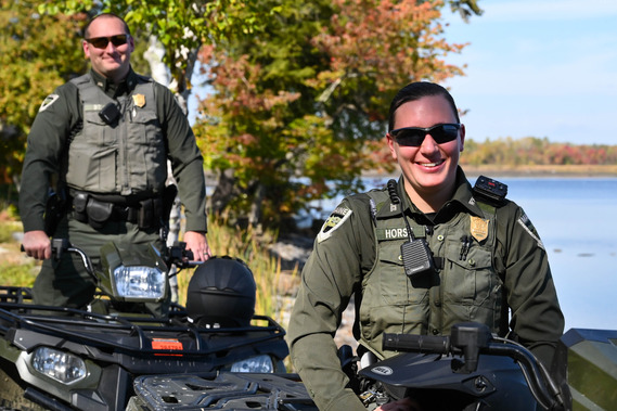 female and male game wardens in green uniforms sitting on ATVs with fall foliage and lake in background