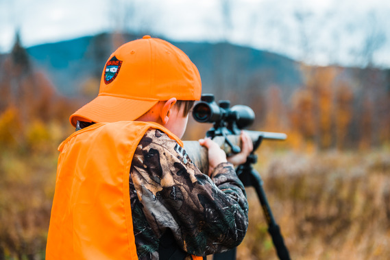 youth hunter wearing two articles of hunter orange shooting a rifle on shooting sticks with fall foliage and mountain background