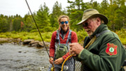 Registered Maine Guide in brown hat and red patch on green sleeve tying a fly for a female fly fishing client on a remote stream