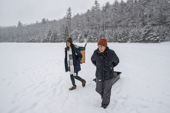 two females carrying ice fishing gear onto ice with fresh snow on trees and on ice