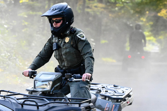 A Maine Game Warden in a green uniform and black helmet riding an ATV on a dusty woodland trail.