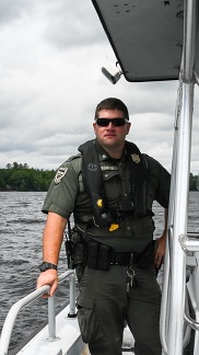 A Maine Game Warden in uniform in a white boat on a lake on a sunny summer day.