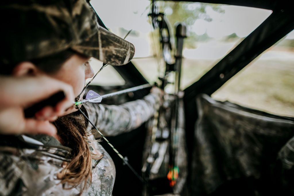 female archer wearing camo in a blind pulling back a bow and arrow