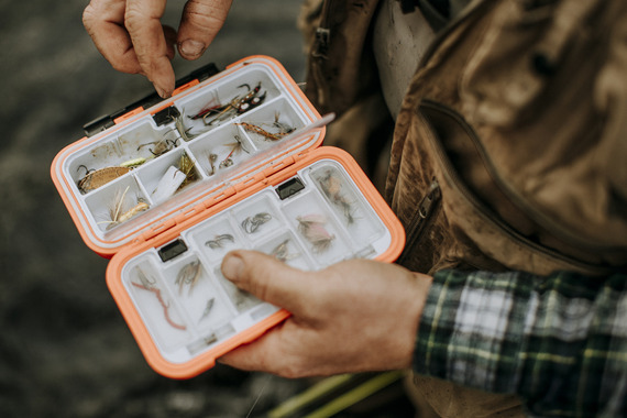 fly angler looking through fly box wearing a tan fly fishing vest and flannel shirt